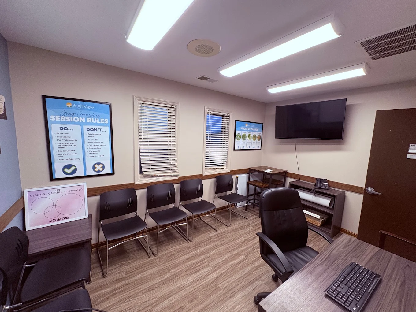 BrightView Health group counseling room with rows of black chairs, wall-mounted TV, counseling posters, windows, workstation, and wood-look flooring
