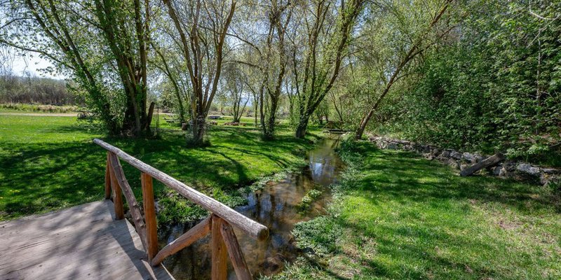 A peaceful outdoor scene with a small wooden bridge crossing a gentle stream surrounded by lush green grass and tall, leafy trees.