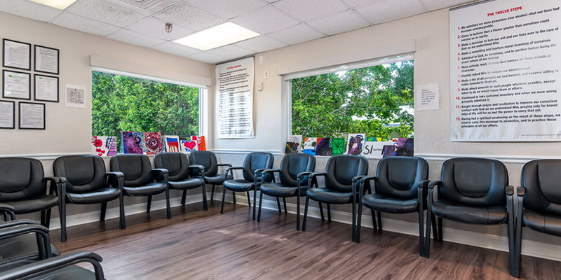 A bright waiting room with black chairs arranged neatly along two walls, large windows providing natural light and a view of greenery, and artwork displayed on the windowsills.