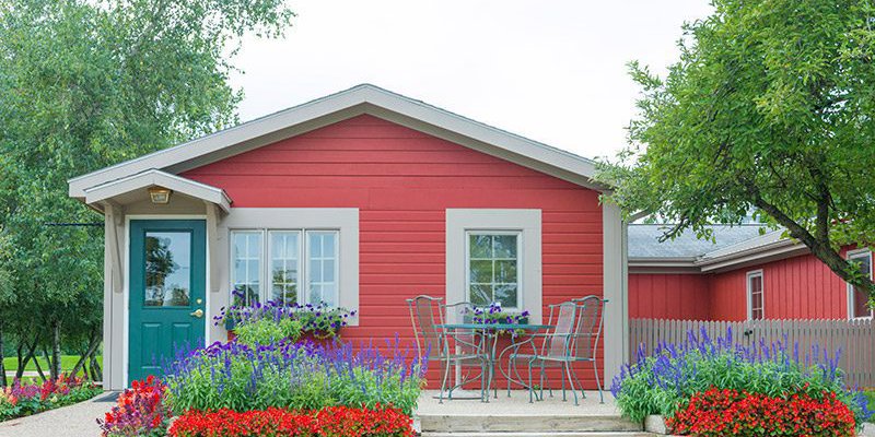 A charming red cottage with a green door is surrounded by vibrant flower beds. A small outdoor seating area adds to the cozy and inviting feel.