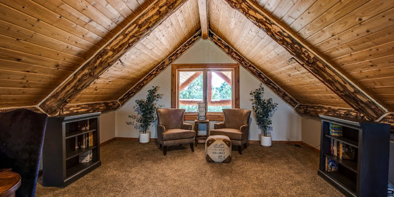 A cozy loft with sloped wooden ceilings, featuring two comfortable chairs by a window, built-in bookshelves, and soft carpeting, perfect for relaxing.