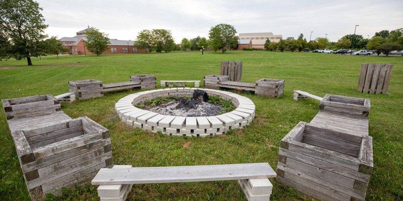  A welcoming fire pit area surrounded by rustic wooden benches and planters, set in a spacious green field, perfect for outdoor gatherings.