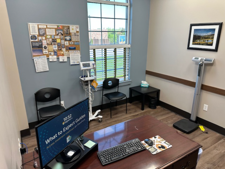 BrightView Health clinical room with desk, computer, patient chairs, vital signs equipment, scale, bulletin board, and window