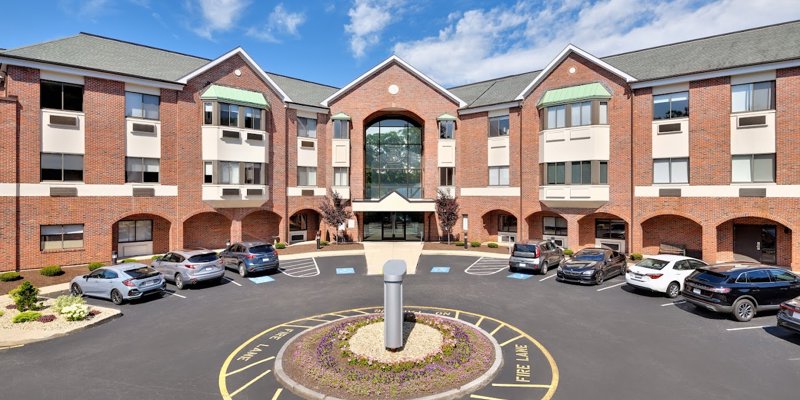  Bright brick building with large windows and a welcoming entrance, surrounded by a neatly landscaped parking area under a clear blue sky.