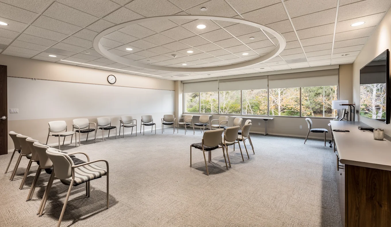 Spacious meeting room with chairs arranged in a circle, large windows, and a wall-mounted television