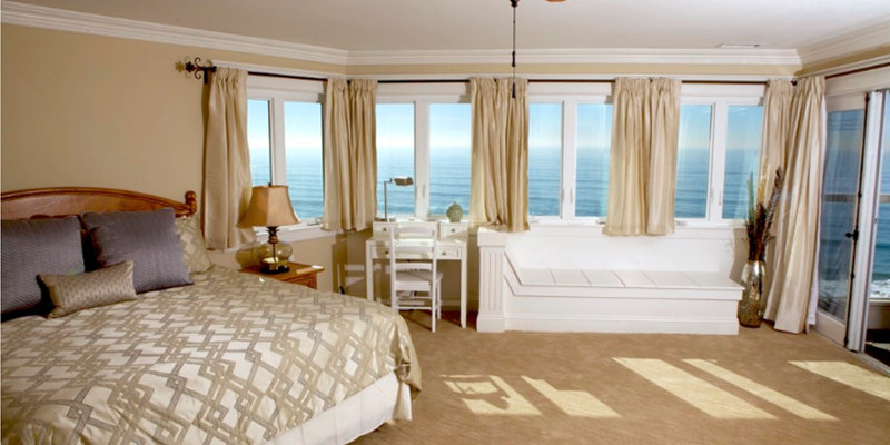  A cozy, sunlit bedroom with ocean views features large windows, elegant curtains, and soft beige tones, creating a relaxing atmosphere.