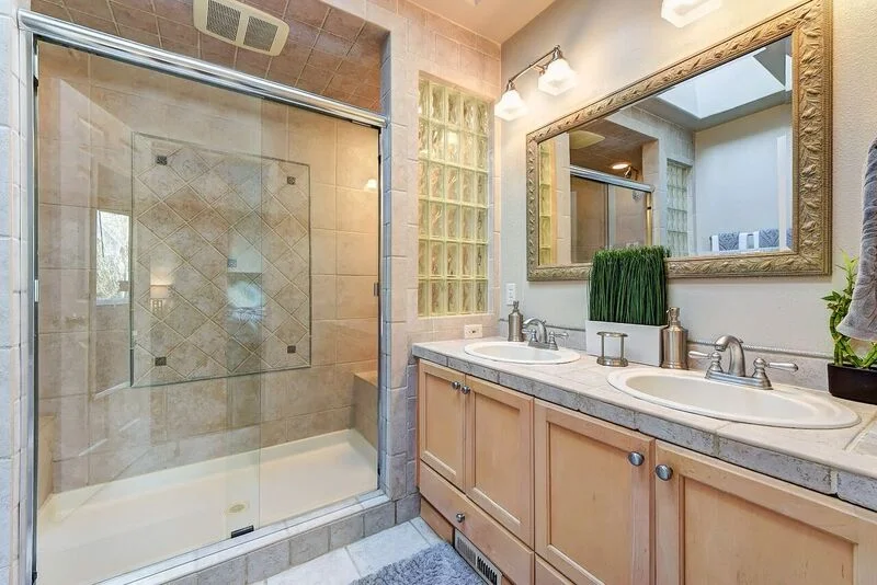 Bathroom with double sink vanity, framed mirror, walk-in glass shower, and beige tile throughout