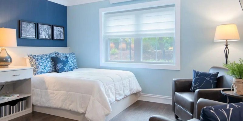 A bright, serene bedroom with soft blue walls, a cozy bed adorned with decorative pillows, and a seating area, creating a peaceful atmosphere.