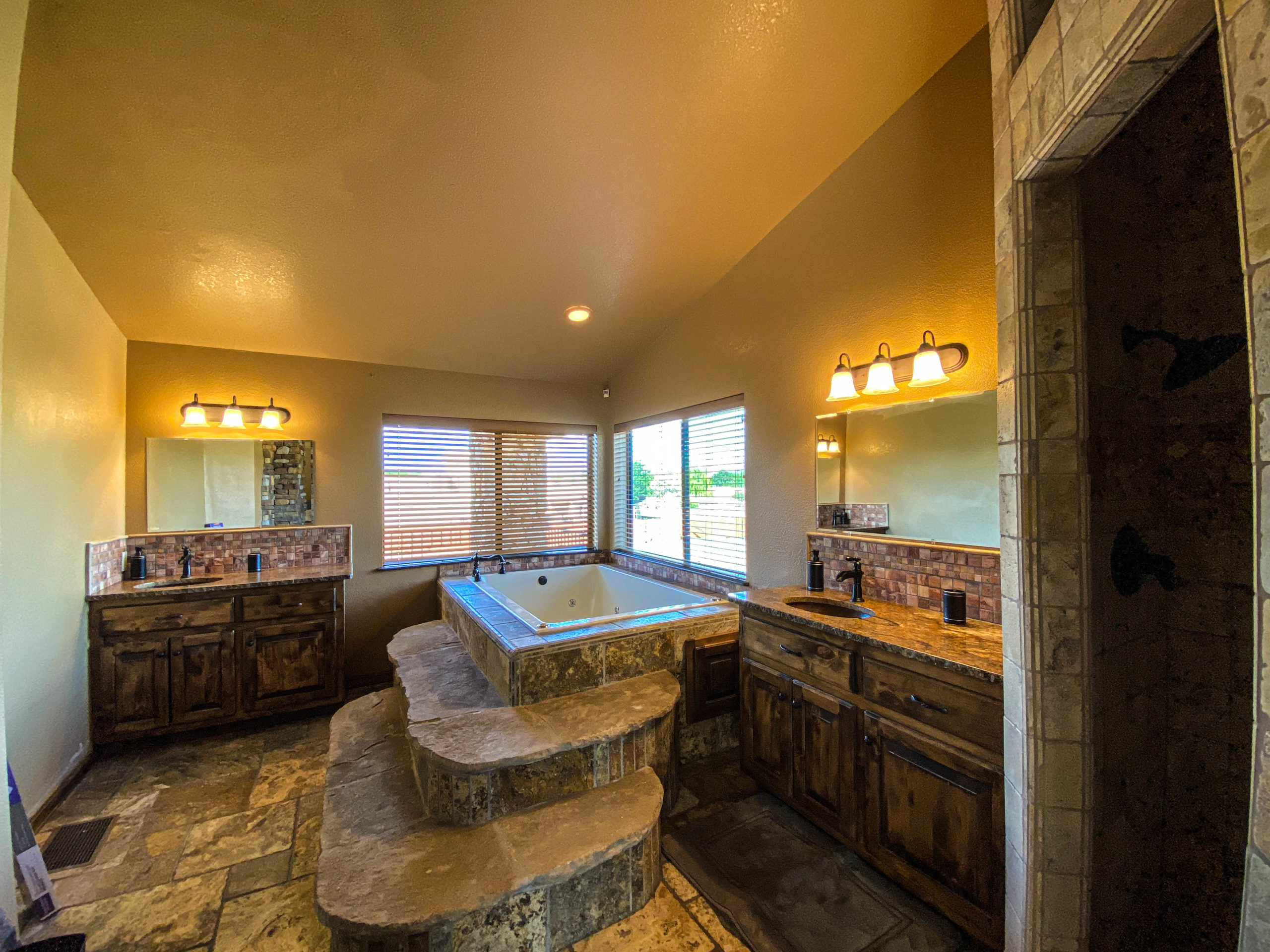 Spacious bathroom featuring a stone step-up tub, double vanities, mirrors, and warm lighting