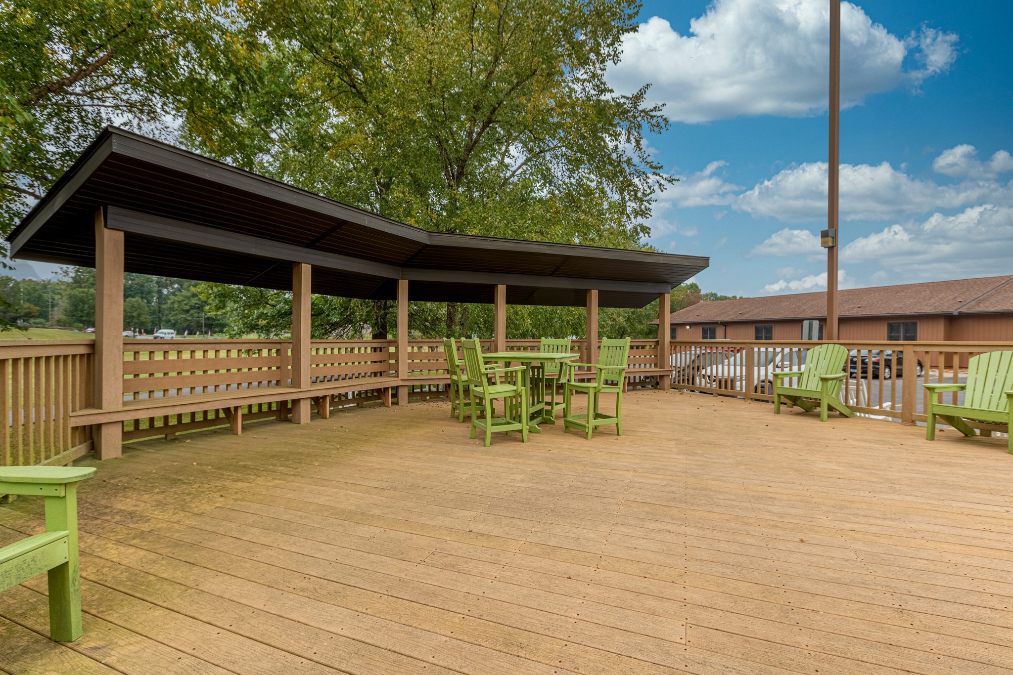 Covered wooden patio with seating, surrounded by trees and facility buildings.