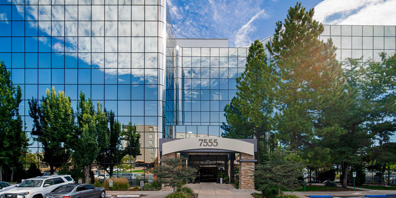 A sleek, mirrored office building at 7555 reflects the blue sky and clouds, surrounded by greenery and well-maintained landscaping with parking in front.