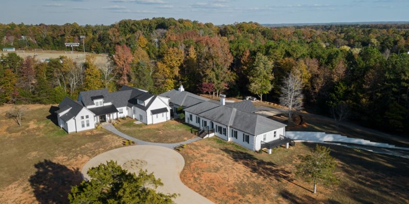 An expansive, elegant estate surrounded by vibrant autumn trees, featuring multiple buildings and a circular driveway in a serene, wooded setting.