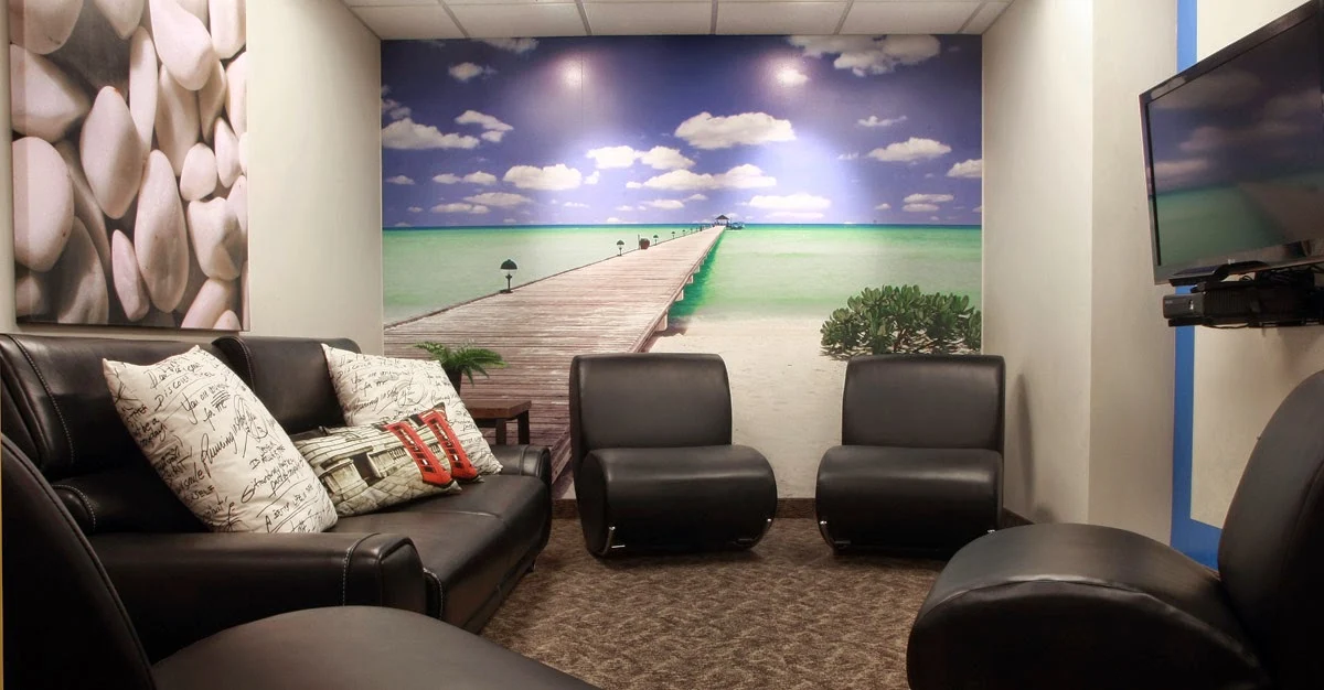 Comfortable lounge with black leather seating, a wall-mounted television, and a large tropical pier mural