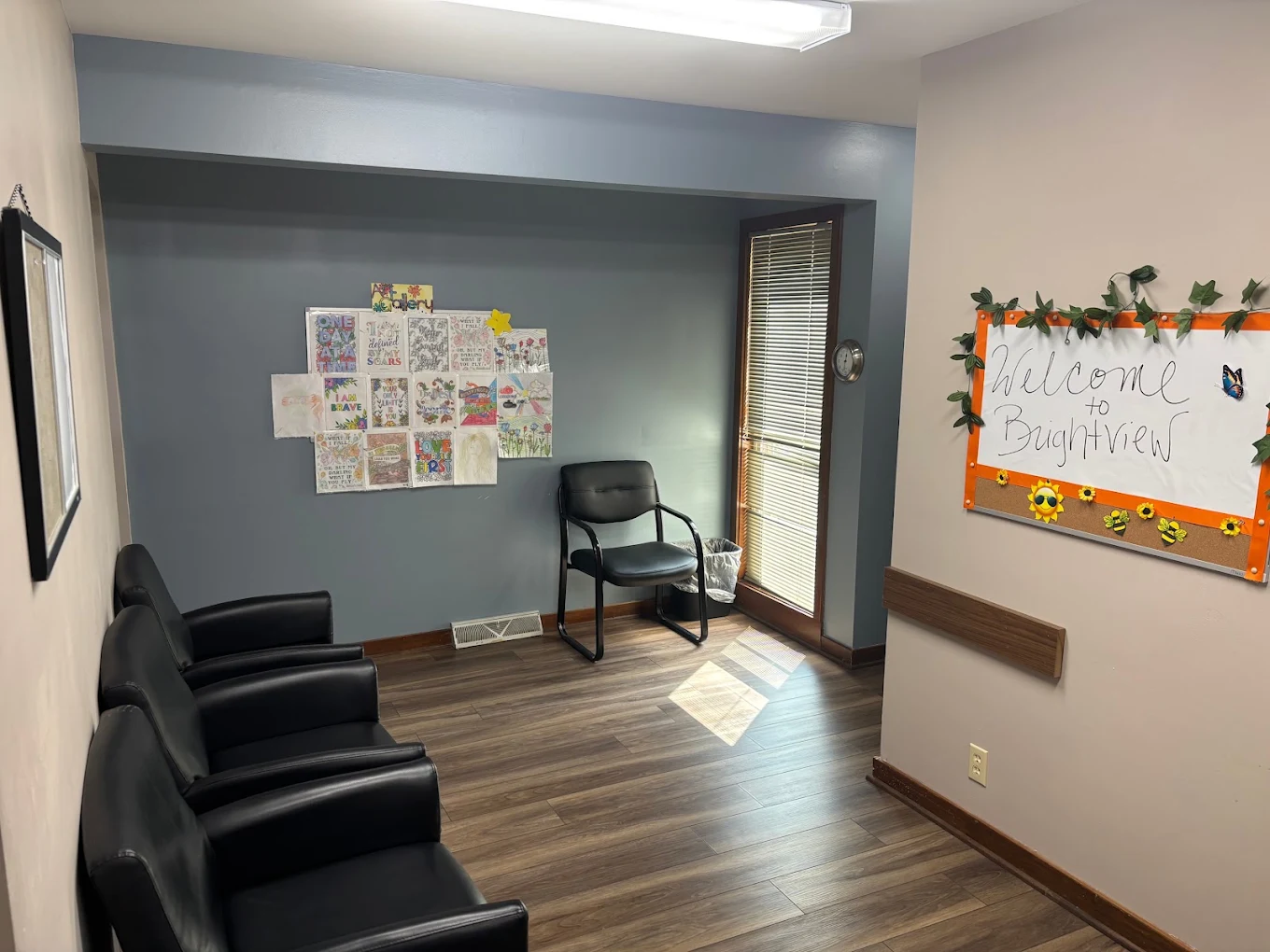 BrightView Health waiting area with black cushioned chairs, blue accent wall, motivational artwork, welcome board, window, and wood-look flooring