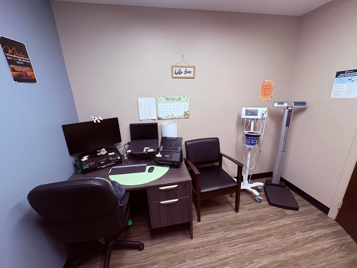 BrightView Health clinical room with desk and computer workstation, patient chair, vital signs monitor, medical scale, wall calendar, and wood-look flooring