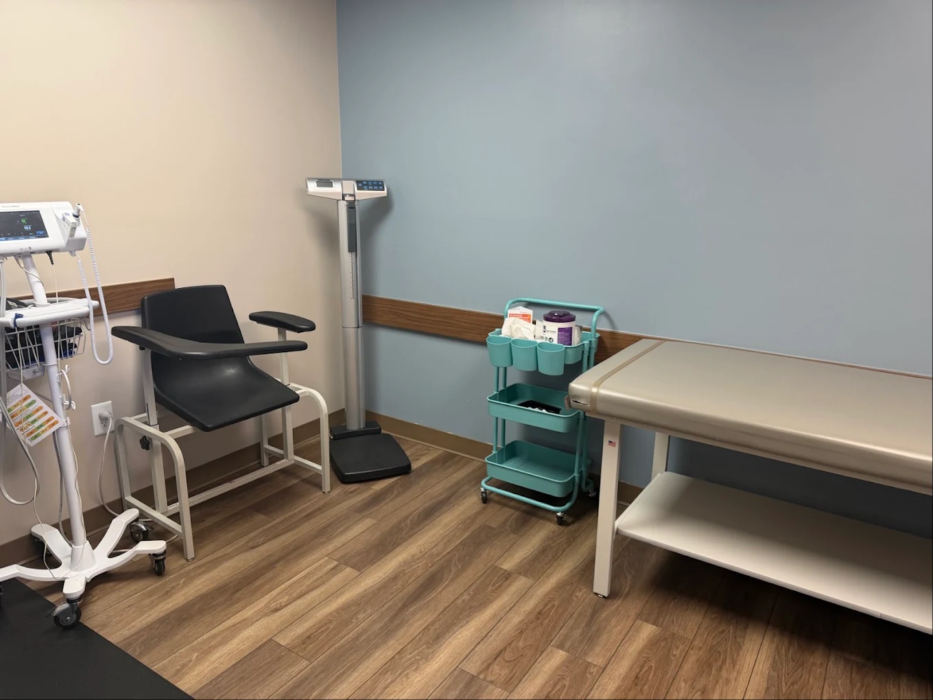 Clinical exam room with patient chair, vital-sign equipment, medical scale, supply cart, and exam table