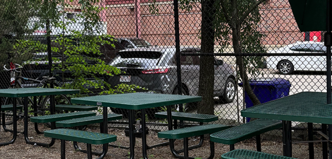 Outdoor picnic tables in fenced area with trees and parked cars in background