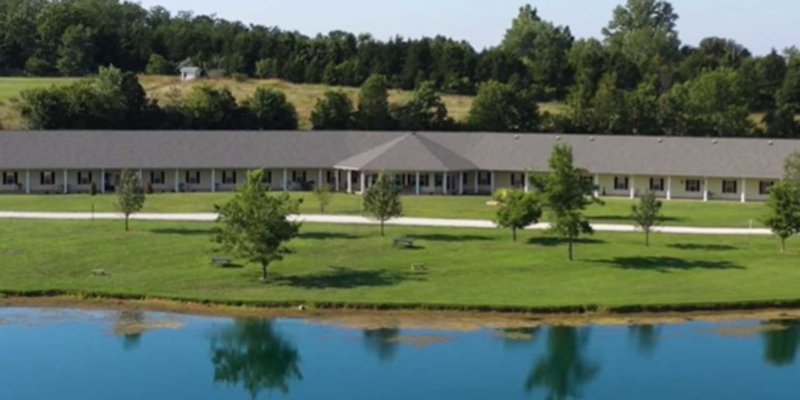 A serene lakeside building, surrounded by lush green grass and trees, reflecting peacefully in the calm water under a bright sky.