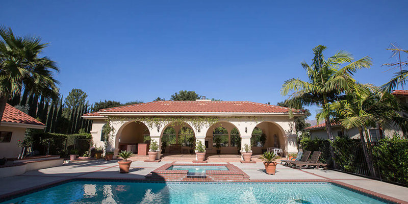 A stunning pool area on a sunny day, featuring a spacious patio with elegant arches, palm trees, and lush greenery, perfect for outdoor relaxation.