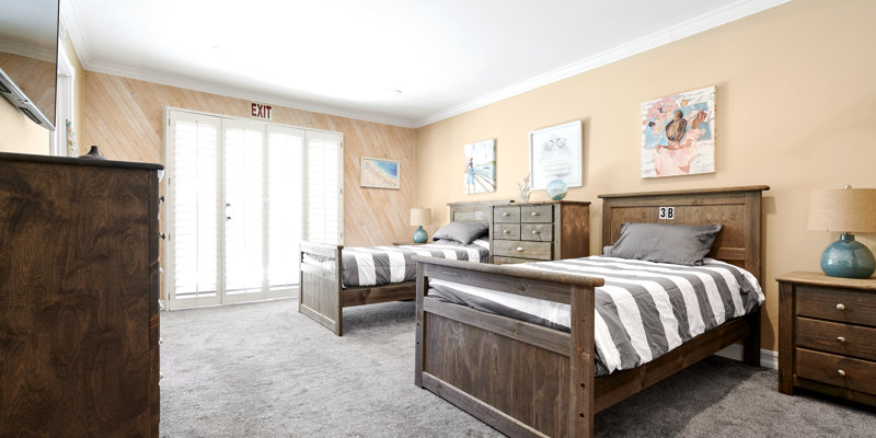 A bright, spacious bedroom with twin beds, cozy striped bedding, and wooden furniture. Natural light streams in through double doors.