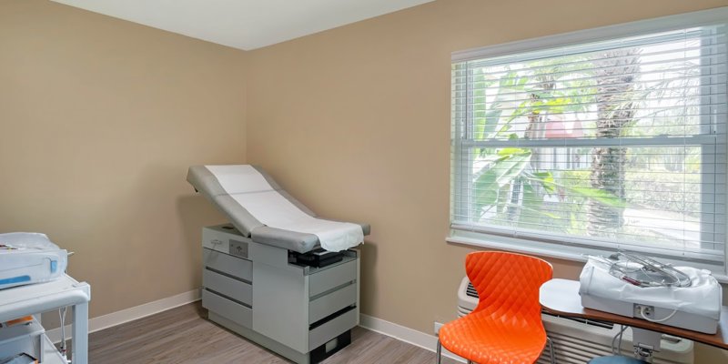 A well-lit medical examination room featuring a comfortable exam table, modern equipment, and a bright orange chair by the window.