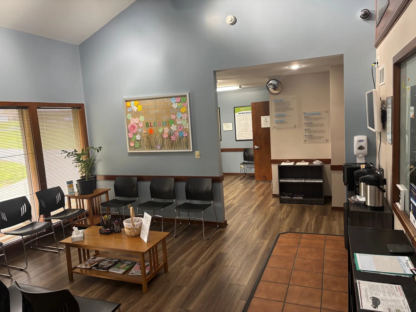 BrightView Health waiting room with rows of black chairs, wooden coffee table, informational materials, beverage station, bulletin board, and wood-look flooring