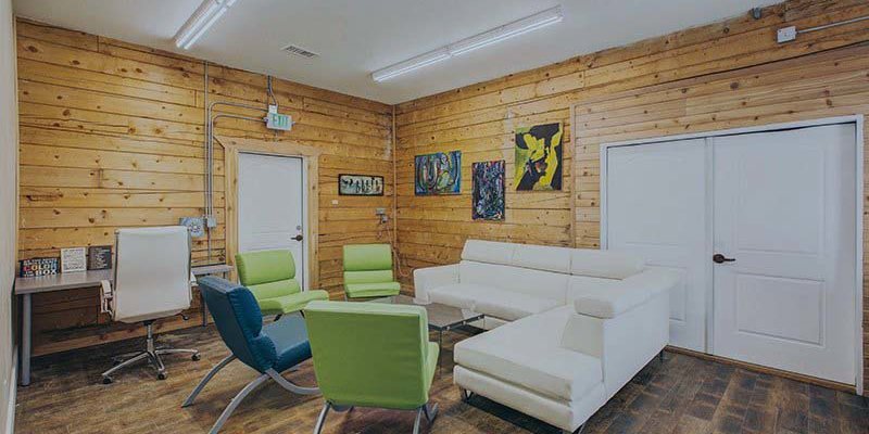 A cozy lounge area with wooden walls, modern white couches, colorful artwork, and vibrant green chairs, creating a warm and inviting atmosphere.