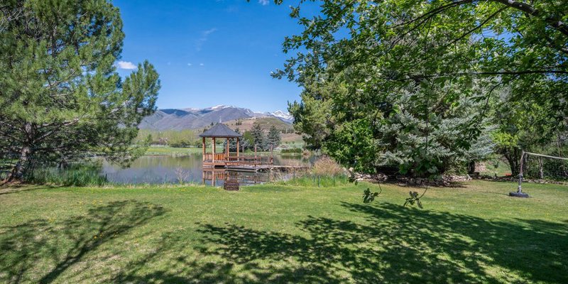 A serene lakeside view with a wooden gazebo surrounded by greenery, mountains in the background, and clear blue skies above. Peaceful and scenic.