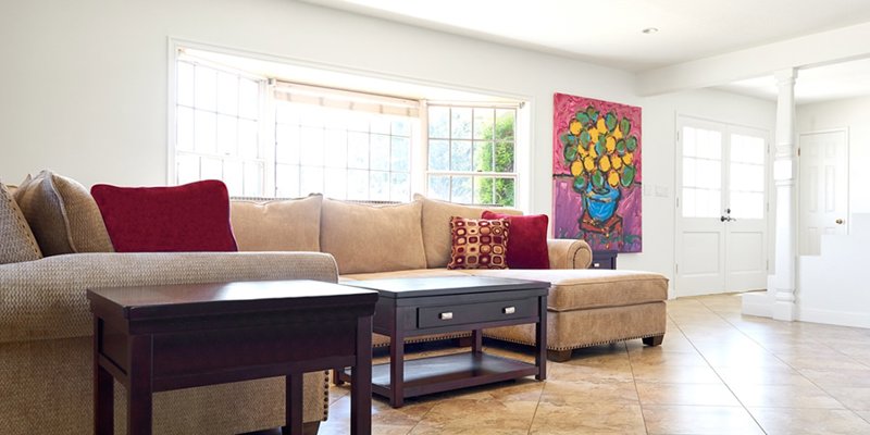 A bright and spacious living room featuring a cozy beige sectional sofa with vibrant red pillows, large windows, and colorful wall art.