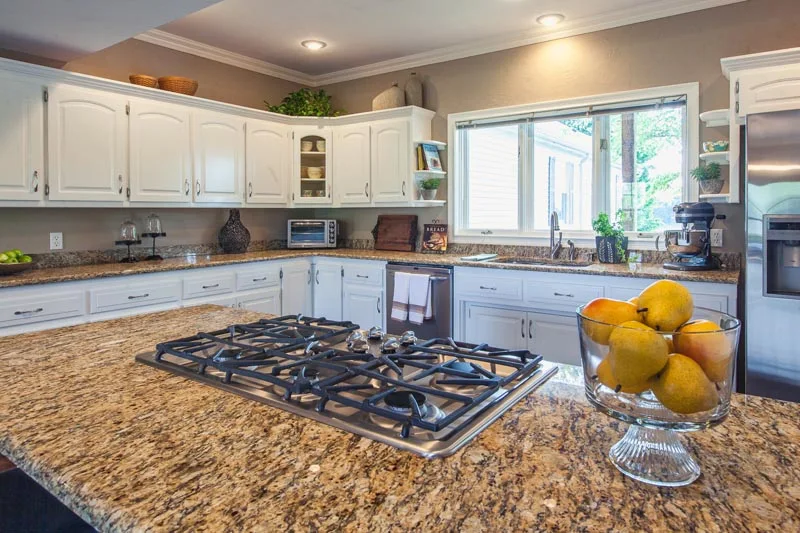 Bright kitchen with granite countertops, gas cooktop island, white cabinetry, and a large window