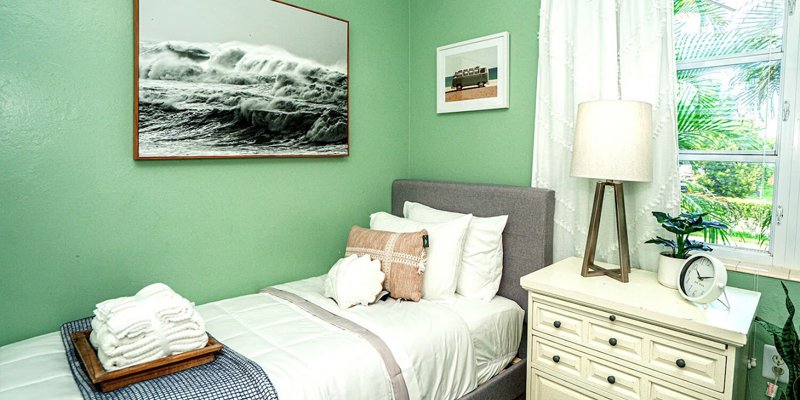 A cozy bedroom with a single bed, soft green walls, and a peaceful ocean-themed painting, creating a serene and comfortable environment.