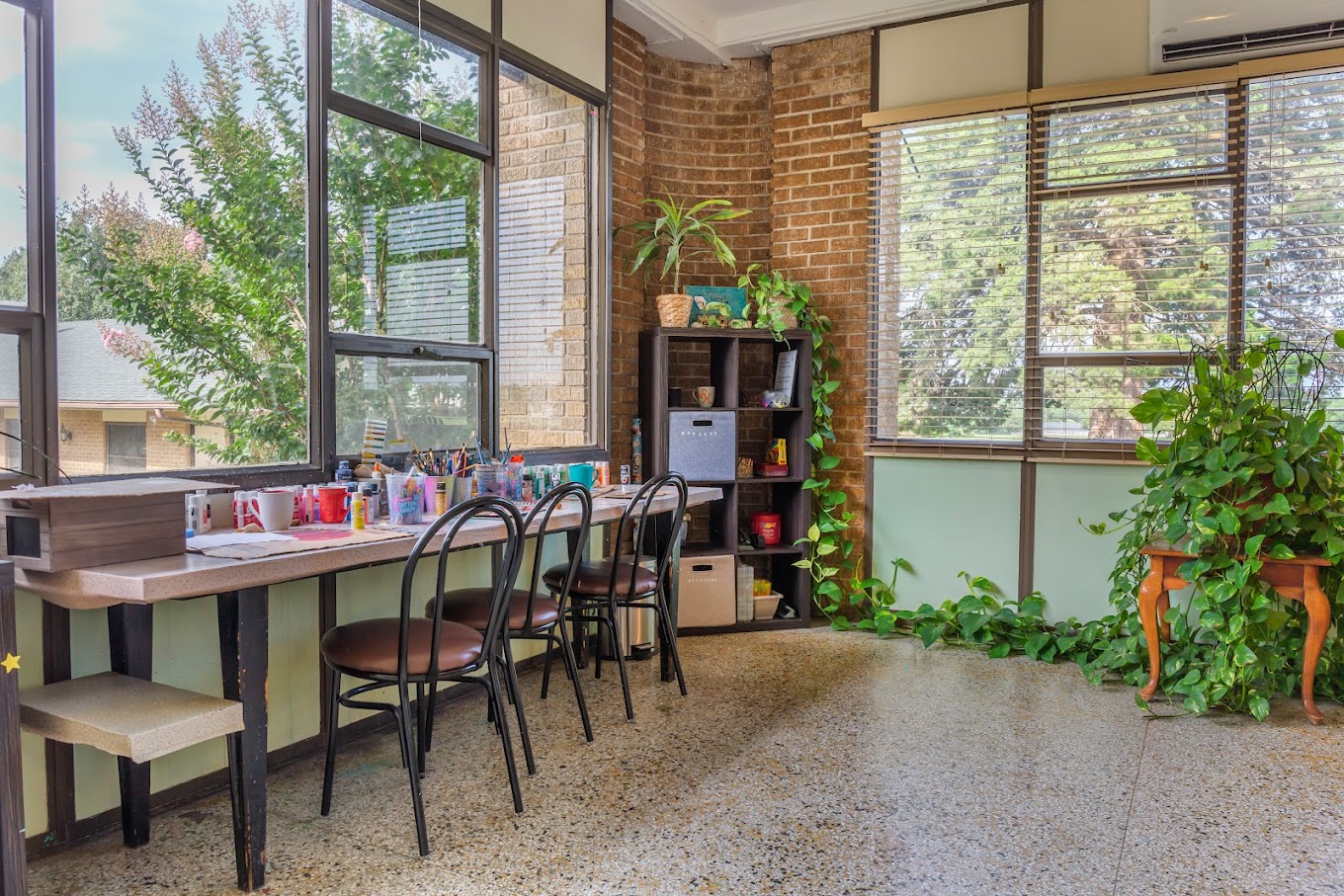 A bright and inviting art room with large windows, a long desk with chairs, and various art supplies, surrounded by greenery.