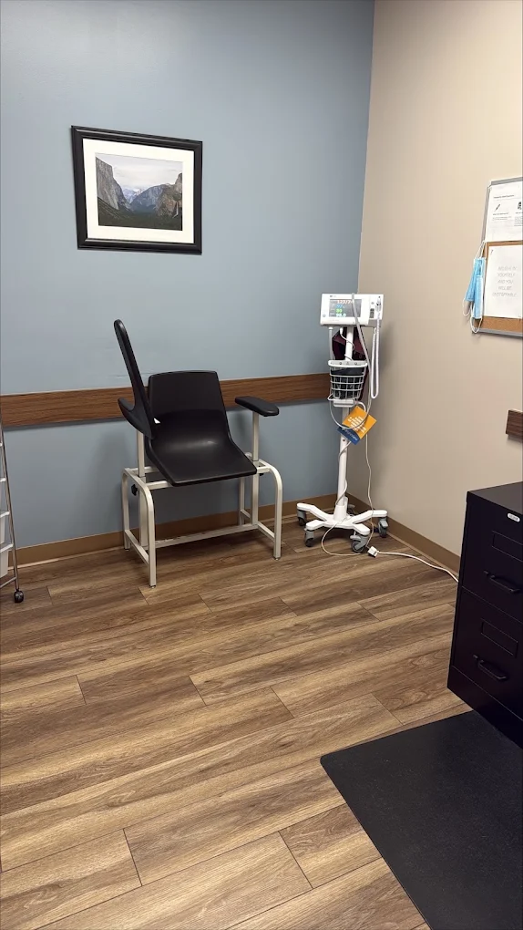 clinical room at BrightView Health Marion with patient examination chair, mobile vital-sign monitoring equipment, blue accent wall, framed landscape artwork, and wood-look flooring