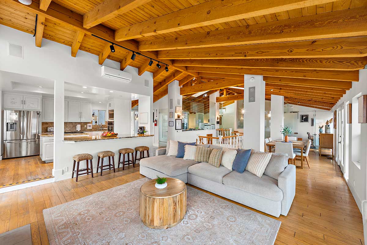 Open living area with large sectional sofa, wood coffee table, vaulted wood-beam ceiling, and adjoining kitchen and dining spaces