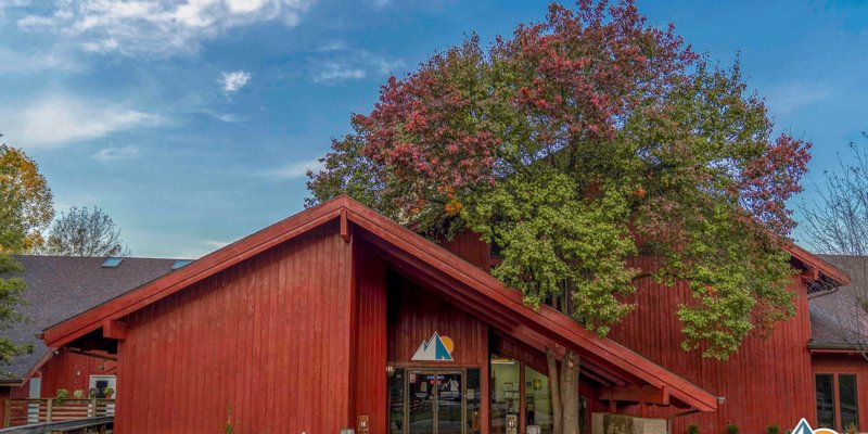 Charming red building surrounded by lush trees with autumn colors, offering a serene and welcoming entrance under a bright blue sky.