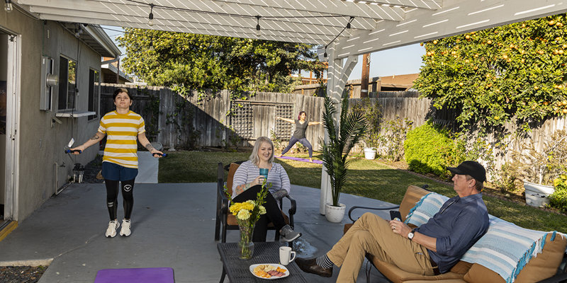 People relax and exercise under a covered patio. One jumps rope, while others enjoy snacks and conversation, creating a lively, sunny space.