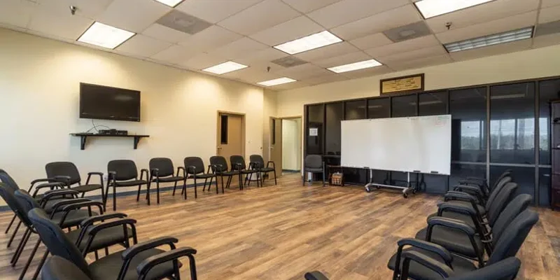 A spacious, well-lit meeting room arranged with chairs in a circle, featuring a whiteboard, TV, and wood-style flooring, ideal for group sessions.