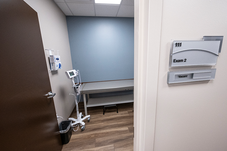 Entrance to an exam room with examination table, mobile vital-sign monitor, medical supplies, and blue accent wall