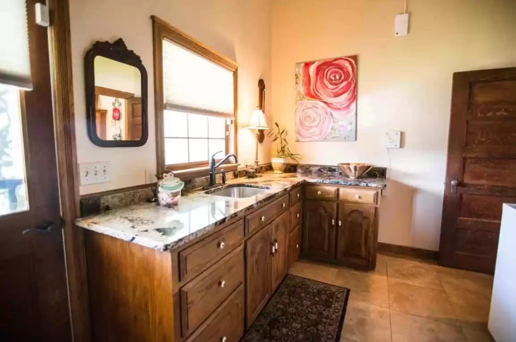 Bright vanity area with granite countertop, wood cabinets, sink, large window, floral wall art, and warm tile flooring