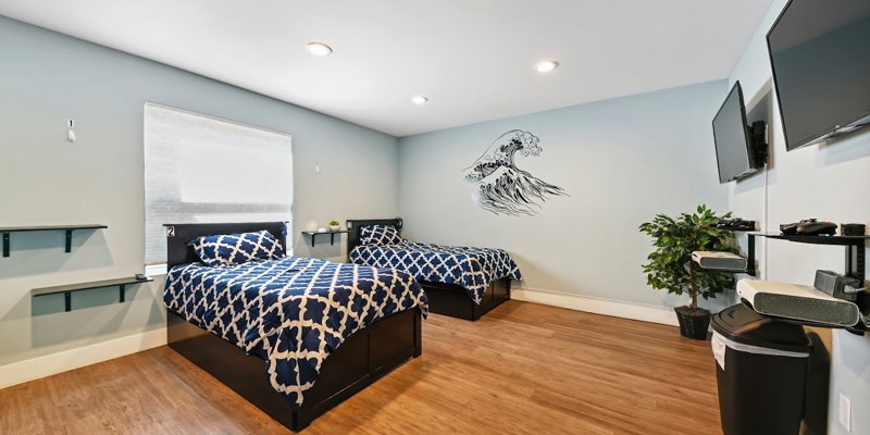 A bright, modern bedroom with two comfortable beds, wall-mounted TVs, and stylish decor, featuring a calming ocean wave mural for a relaxing vibe.