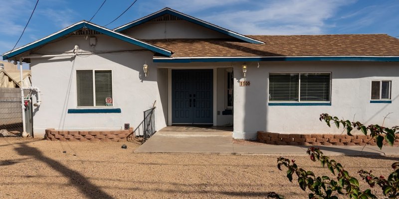  A welcoming single-story home with a blue-trimmed roof and windows, a neat gravel yard, and a large front entrance under a clear sky.