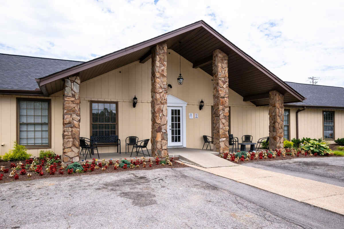 Exterior of a rehab facility with stone columns, a sitting area with black chairs, and landscaping