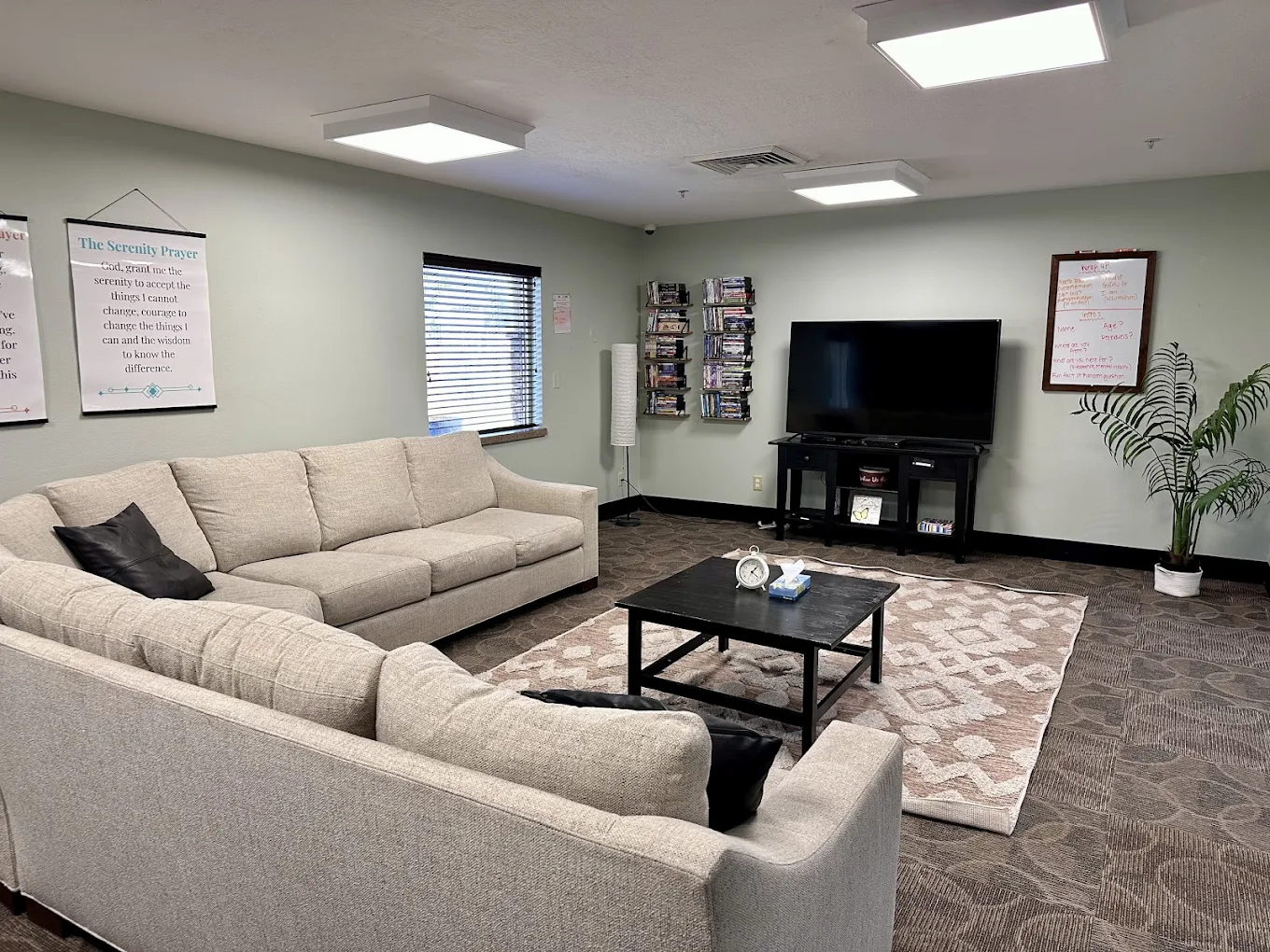 Cozy sitting area with beige couches, coffee table, wall posters, flat-screen TV, and bookshelf at New Roads Behavioral Health