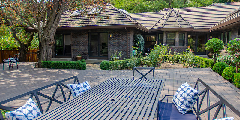 A peaceful outdoor patio with comfortable seating, decorative pillows, and a large table, surrounded by lush greenery and charming brick buildings.