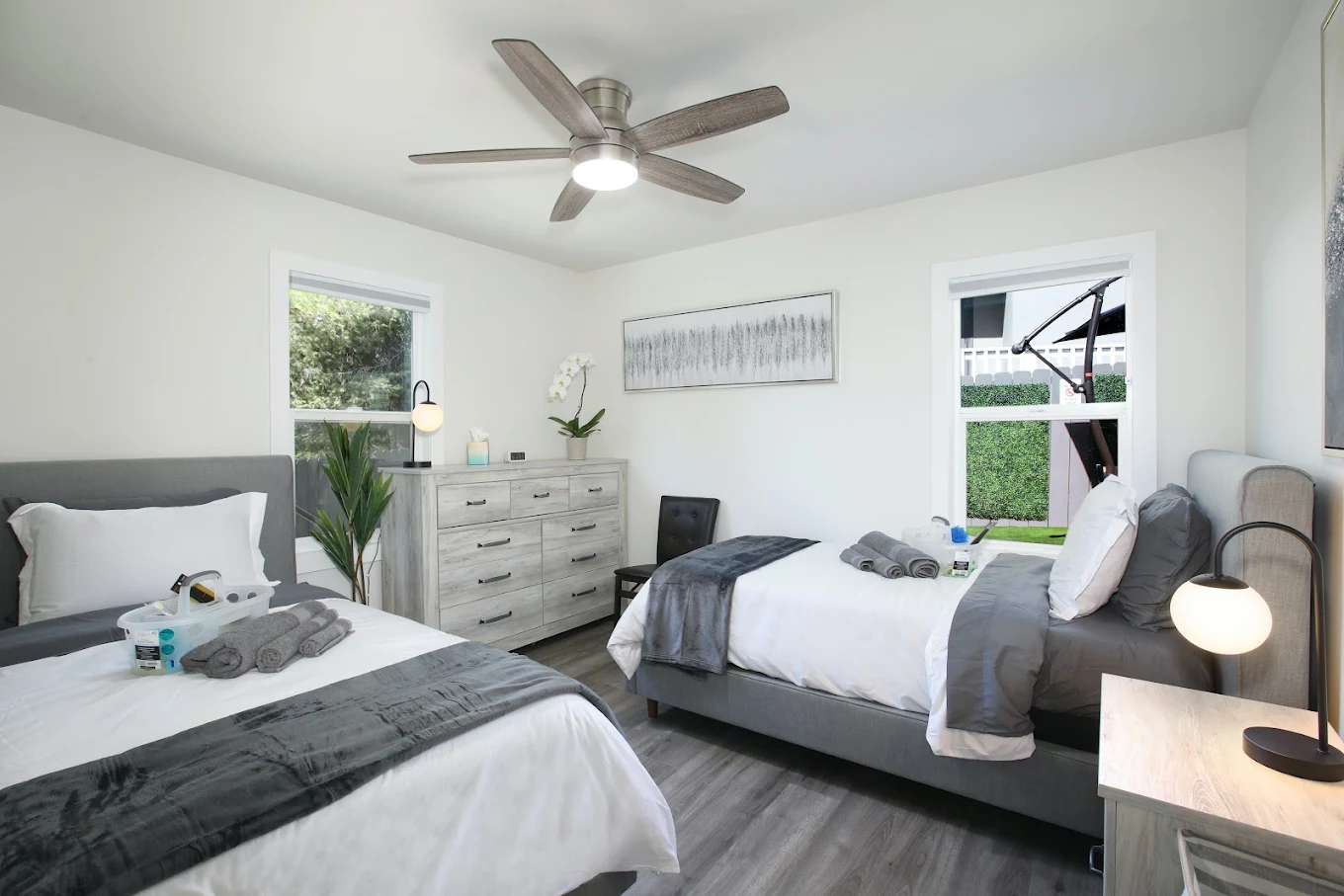 Bright shared bedroom with two beds, gray-and-white bedding, dresser, bedside lamps, and large windows