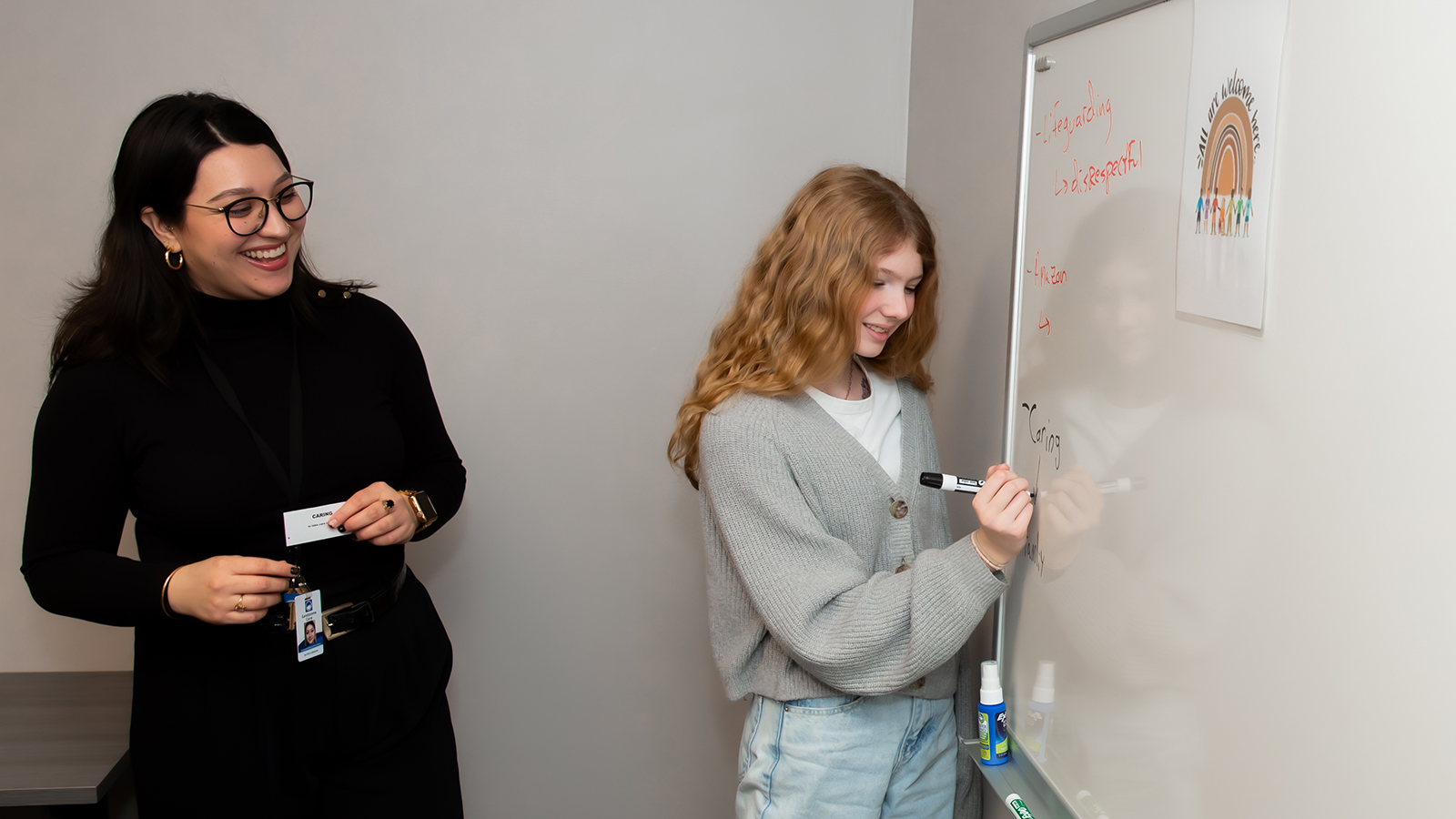 A young woman writes on a whiteboard while another person observes.