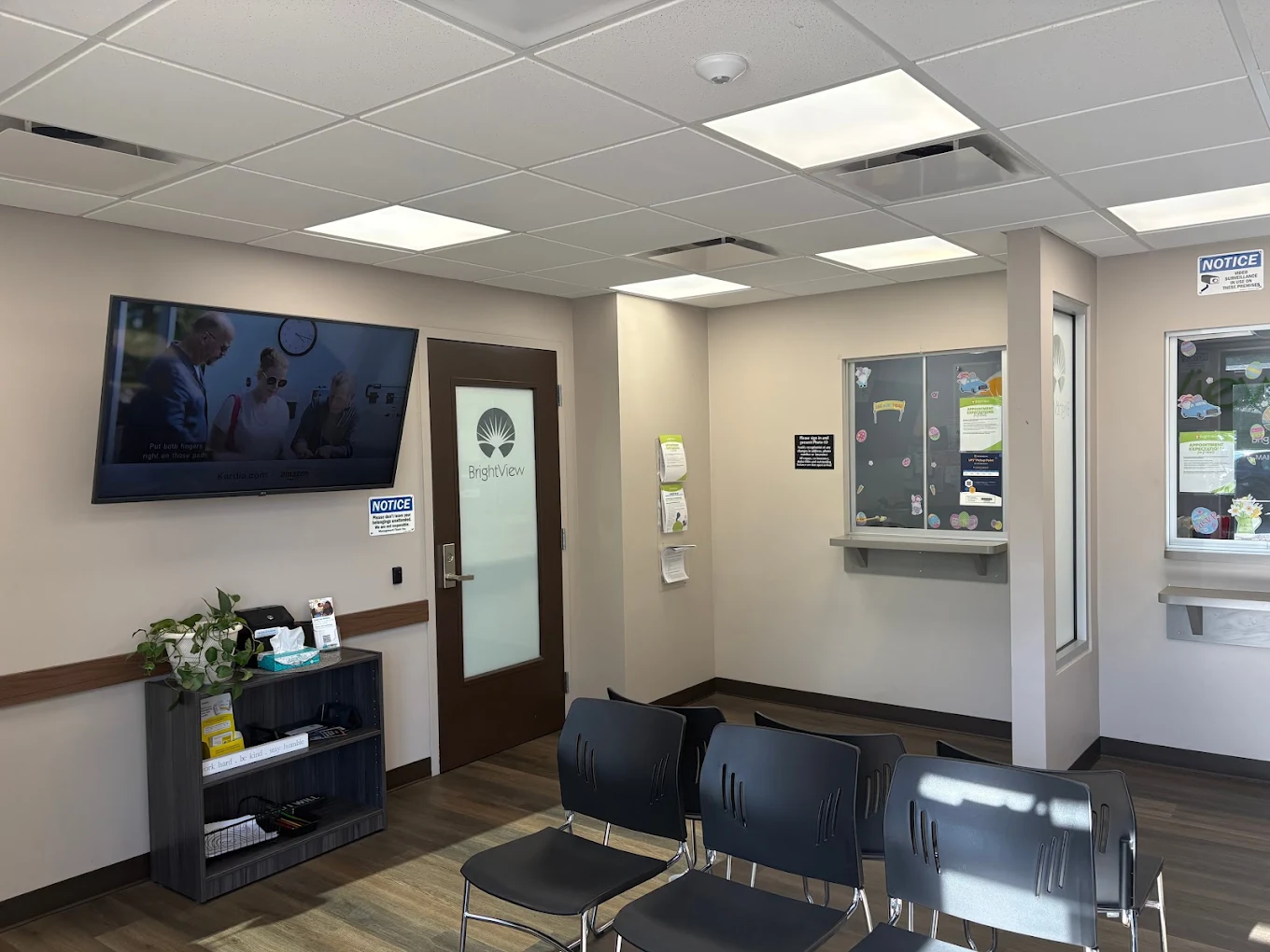 BrightView Health waiting area with rows of black chairs, wall-mounted television, service windows, informational displays, and wood-look flooring