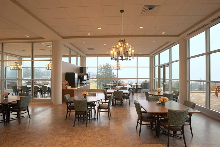 Bright dining area with multiple tables, upholstered chairs, chandeliers, and large windows overlooking the grounds