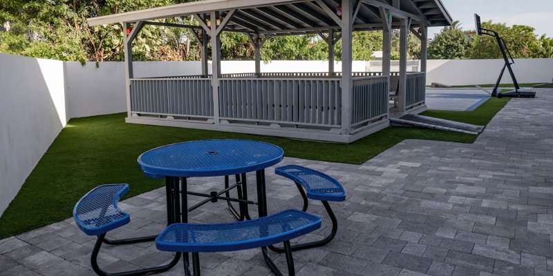 A peaceful outdoor area with a shaded gazebo, blue picnic table, and basketball court.