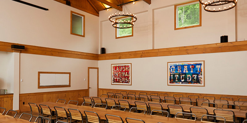 A spacious and rustic meeting hall featuring long wooden tables, modern chairs, large ceiling fans, and decorative lighting, ideal for gatherings.